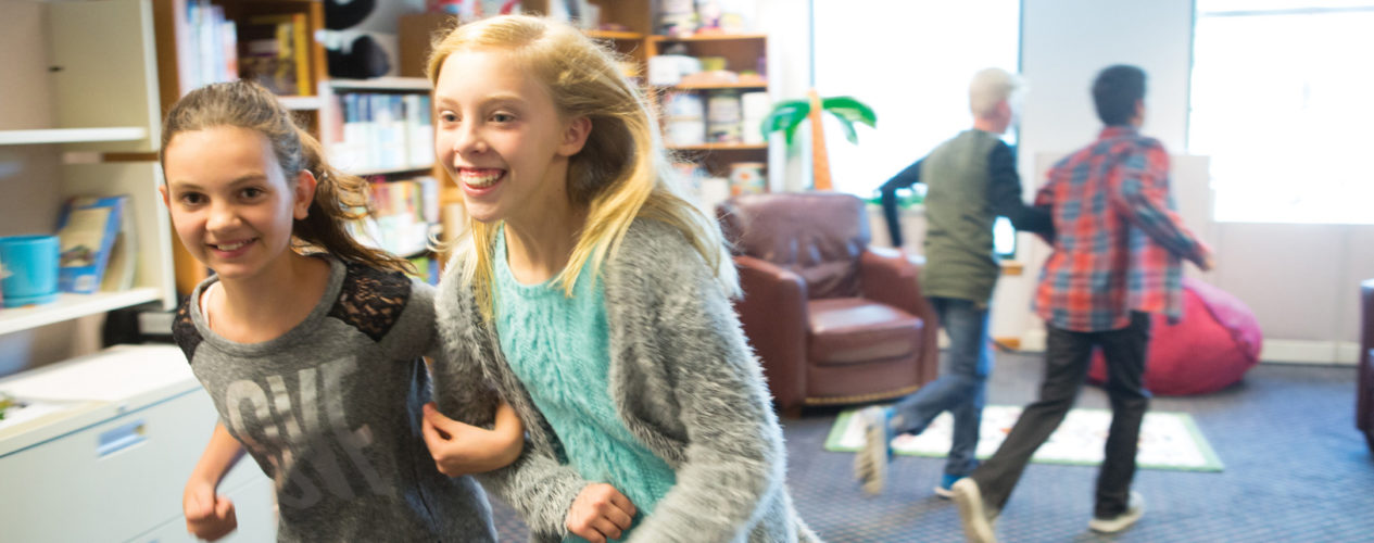Two preteen girls linked arm in arm running around the classroom during a preteen Bible activity.