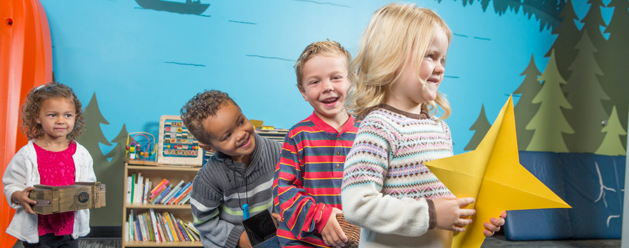 A little girl is holding a gold star made out of construction paper. There are two little boys and a little girl in a line behind her, all smiling. They are in a Sunday school classroom with trees and a lake painted on the walls.