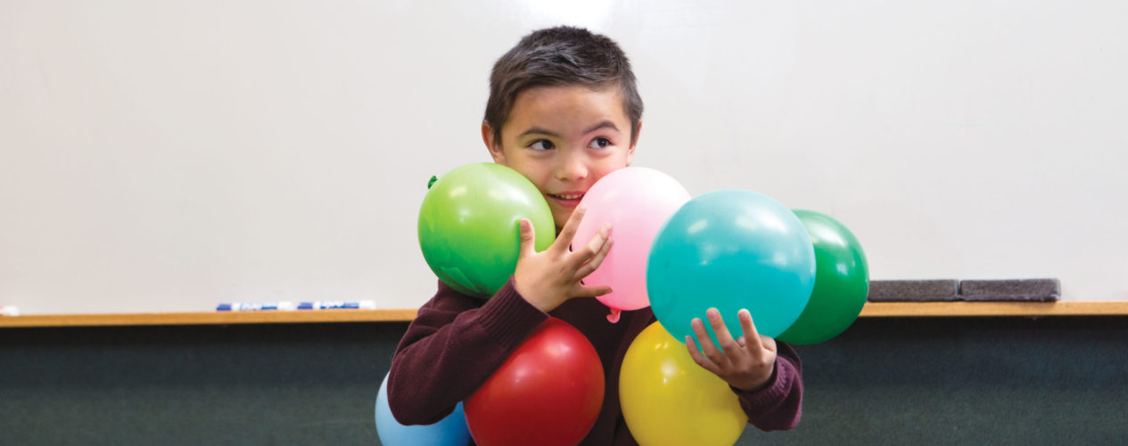 Elementary aged boy trying to hold lots of colorful balloons in his arms.