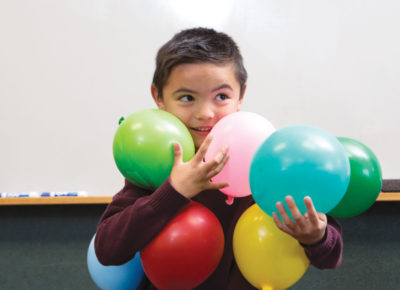 Elementary aged boy trying to hold lots of colorful balloons in his arms.