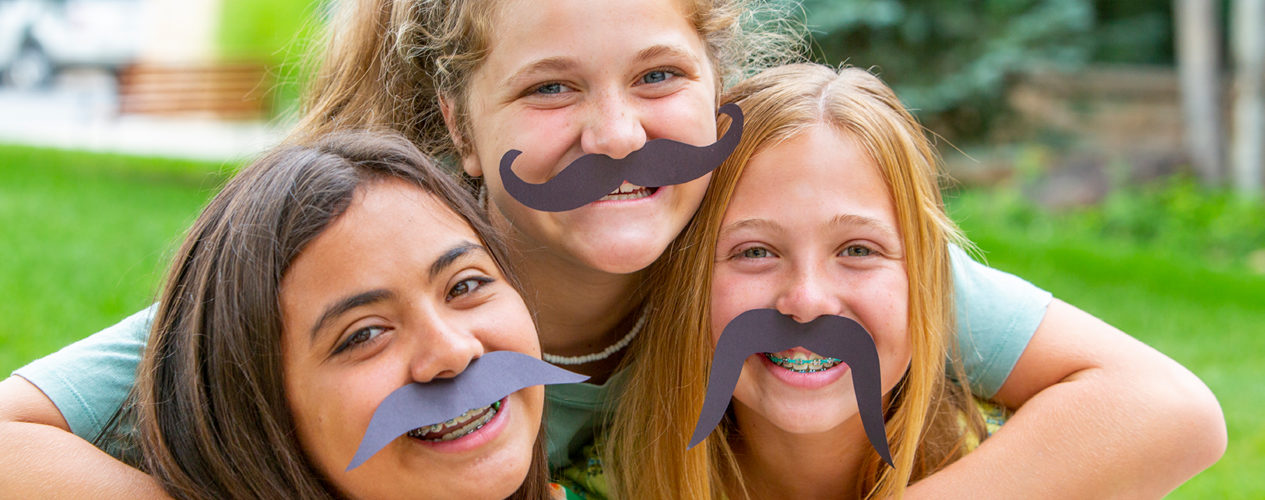Three preteen girls wearing construction paper mustaches.